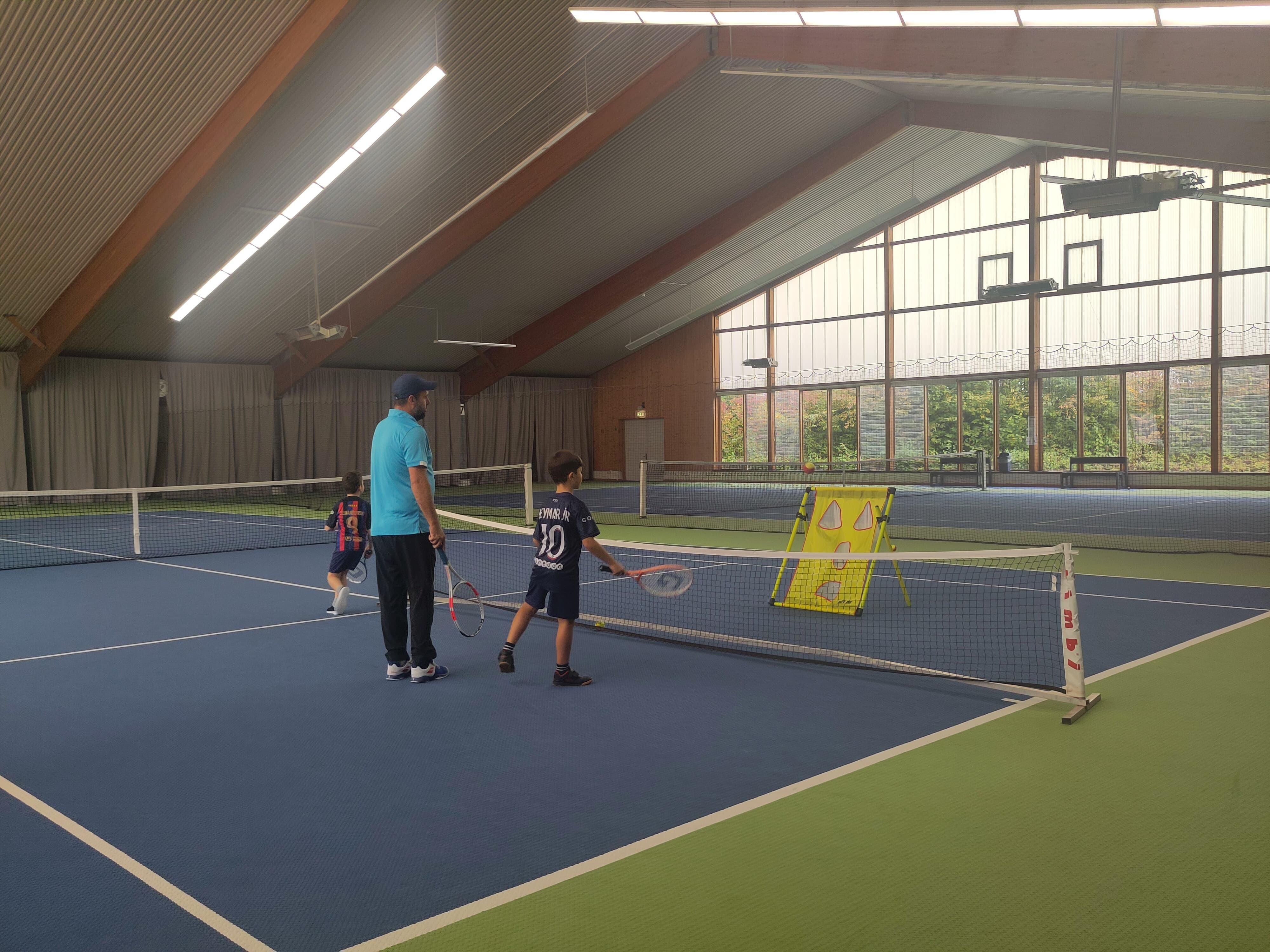 Bildbeschreibung: Kinder stehen in einer Tennishalle um den Trainer. Er erklärt etwas.