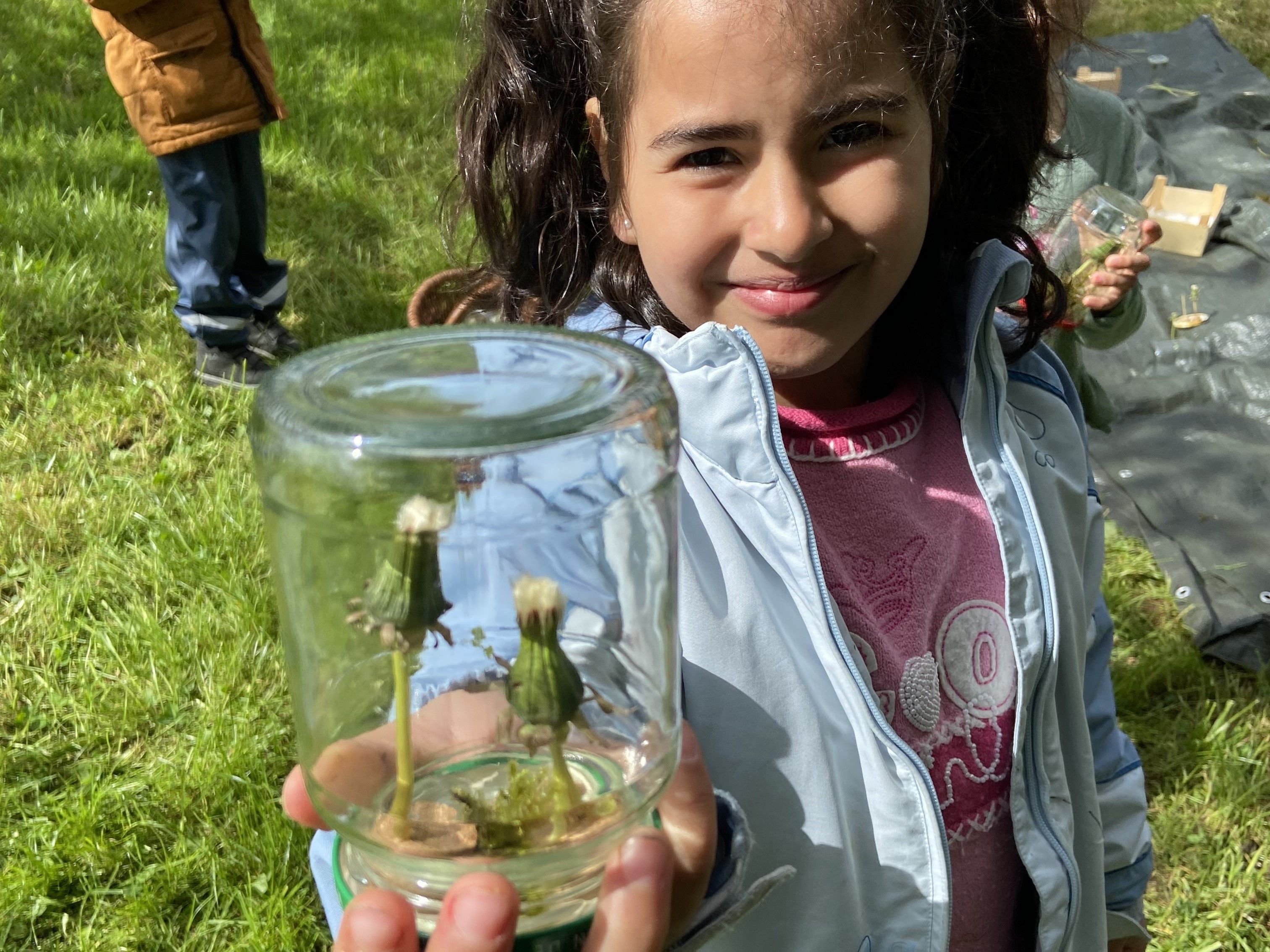 Bildbeschreibung: Kind hält ein Glas mit Löwenzähnen in die Kamera.