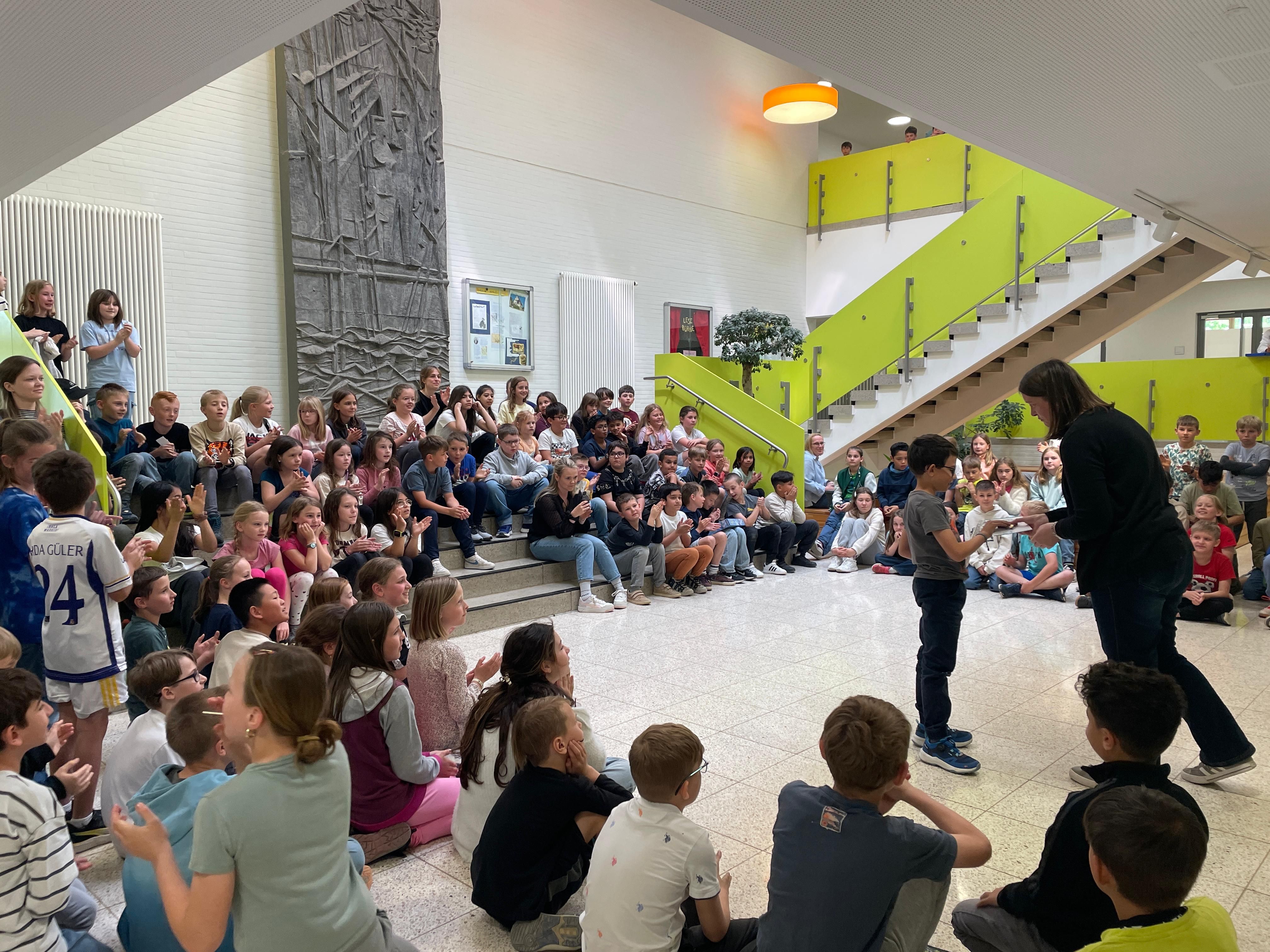 Viele Kinder sitzen in der Aula. Eine Lehrerin überreicht einem Schüler einen Preis.
